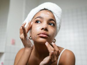 A young woman with a towel wrapped around her head applying facial cleanser in a bathroom.