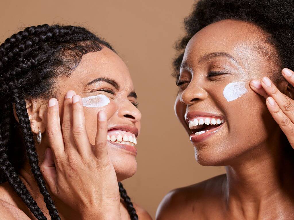 Two women with glowing skin laughing while applying sunscreen to their faces.