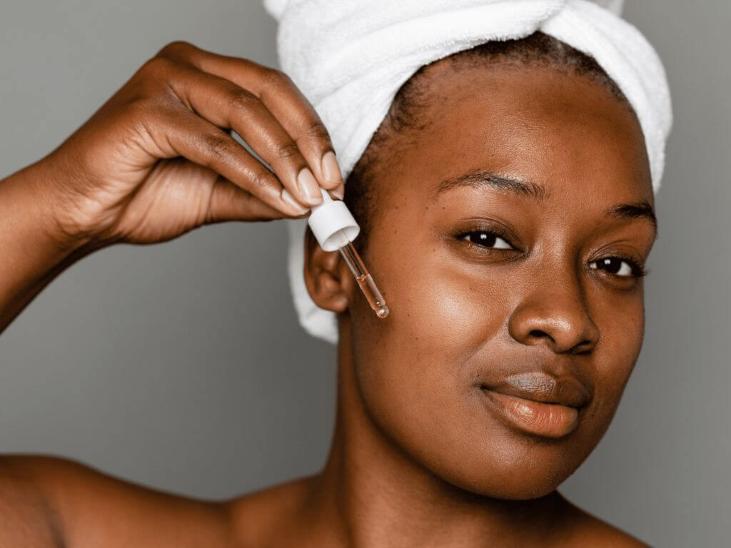 A woman with a towel on her head applying a skincare serum using a dropper.